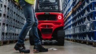 Mit Lösungen von Linde können Sie die Sicherheit von Fußgängern in Ihrem Unternehmen erhöhen. Eine Person in Warnweste läuft an einem Regalgang vorbei, aus dem gerade ein Gabelstapler rückfährts herausfährt.